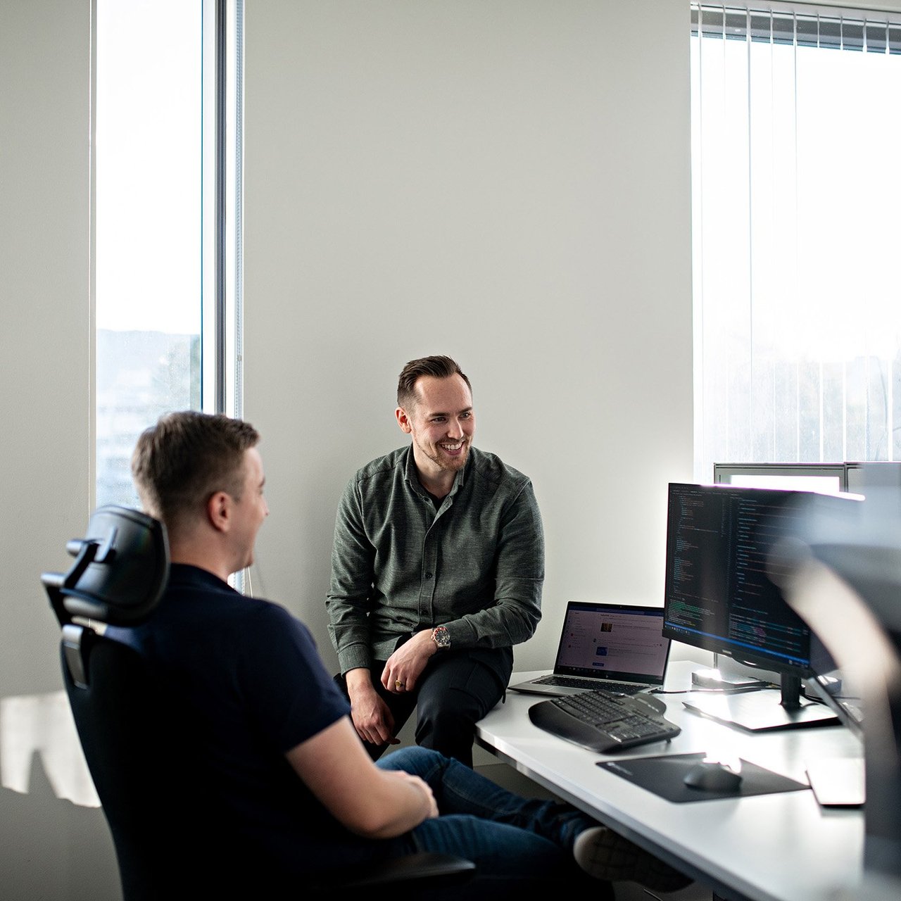 En mann sitter på kontorplassen sin og ser på PC-skjermen med en kollega sittende ved siden av på pulten