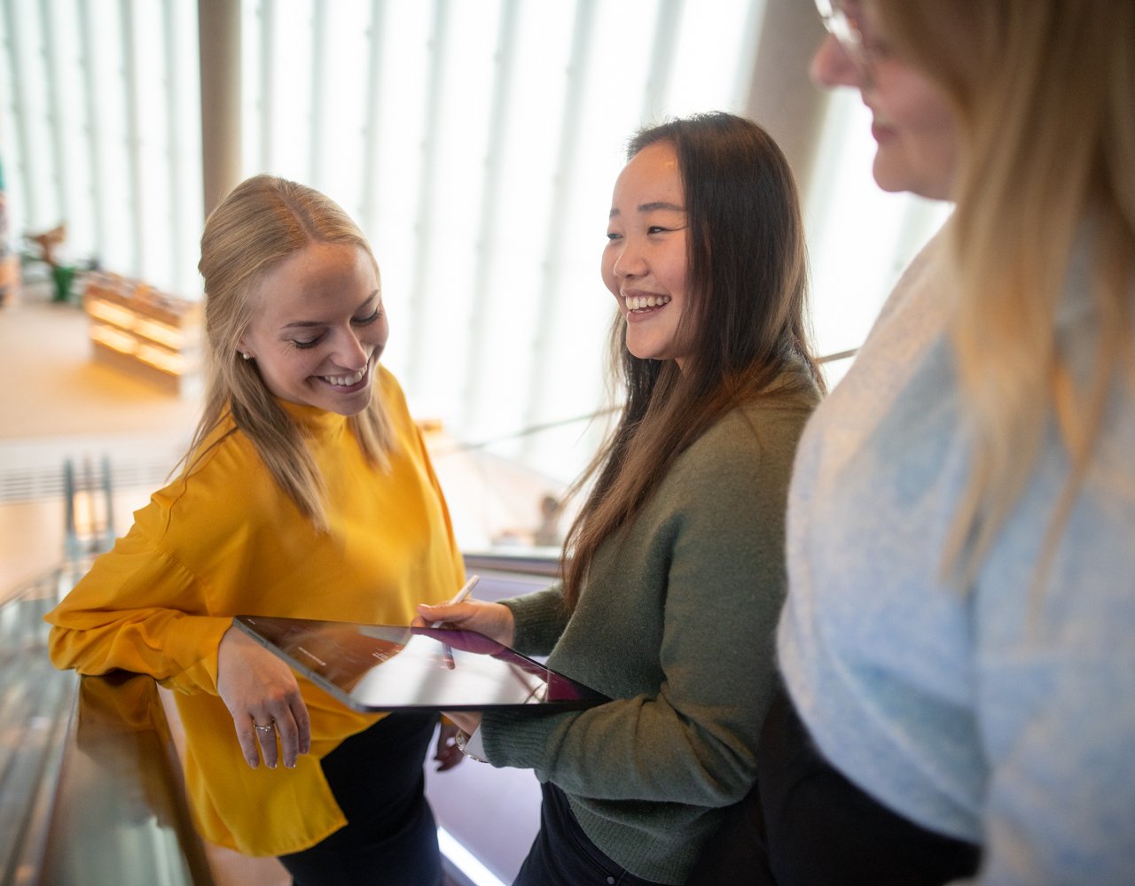 Tre damer står og ler sammen med en ipad og ser på datavisualisering