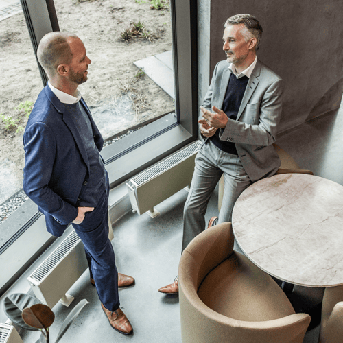 To menn står og snakker med hverandre ved et vindu