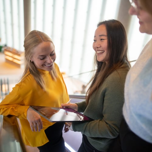 Tre damer står og ler sammen med en ipad i en rulletrapp