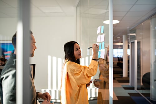 Rådgiver i gul skjorte snakker mens hun skriver på post-it-lapper på glassveggen