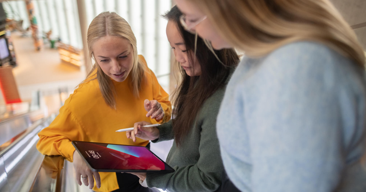 Tre damer stor i en rulletrapp og ser ned på en ipad